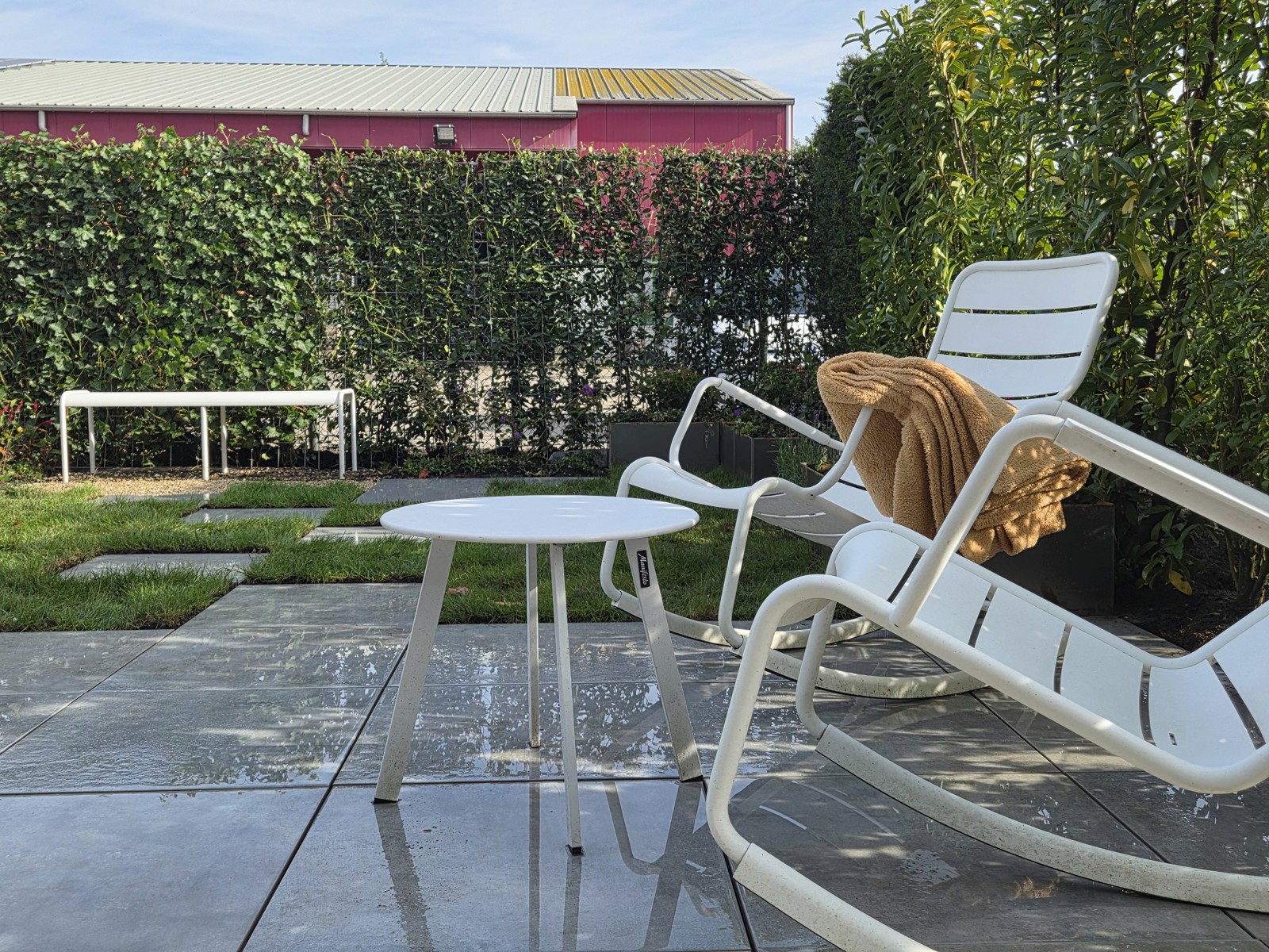 Seating area with white garden chairs and a round table on the terrace next to grass and stepping stones